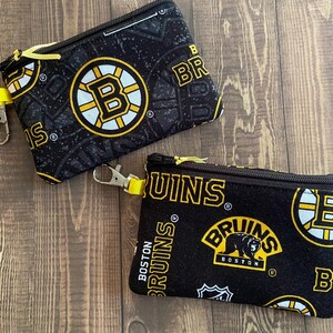 May include: Two black zippered pouches with yellow accents and Boston Bruins logos. The pouches have a black background with yellow and white Bruins logos and text. Each pouch has a yellow zip pull and a silver-coloured clip.