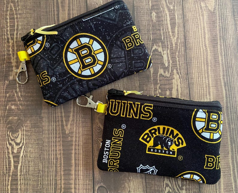 May include: Two black zippered pouches with yellow accents and Boston Bruins logos. The pouches have a black background with yellow and white Bruins logos and text. Each pouch has a yellow zip pull and a silver-coloured clip.