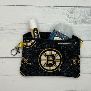 May include: Black zippered pouch featuring the Boston Bruins logo and pattern. The pouch contains a ChapStick and a hand sanitizer bottle. The pouch has a yellow strap and a silver clasp. The background is a white wooden surface.