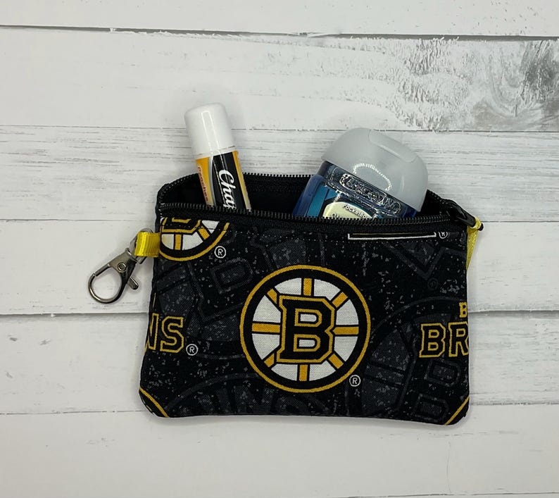 May include: Black zippered pouch featuring the Boston Bruins logo and pattern. The pouch contains a ChapStick and a hand sanitizer bottle. The pouch has a yellow strap and a silver clasp. The background is a white wooden surface.