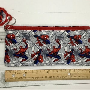 May include: A rectangular fabric pencil case featuring a repeating Spider-Man pattern on a gray web background with a red zipper. A matching octagonal keychain is attached by a silver ball chain. A wooden ruler shows the case is 9 inches long.