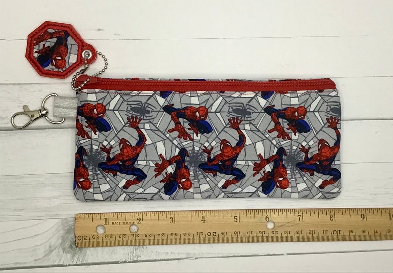 May include: A rectangular fabric pencil case featuring a repeating Spider-Man pattern on a gray web background with a red zipper. A matching octagonal keychain is attached by a silver ball chain. A wooden ruler shows the case is 9 inches long.