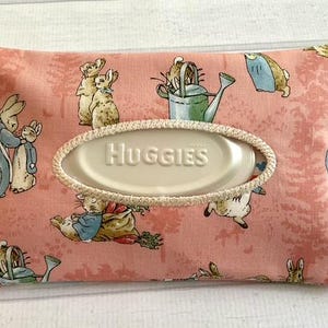 May include: A rectangular, pink fabric tissue holder with a repeating pattern of rabbits and garden tools. The holder has an oval opening with the word "Huggies" and a cream-colored trim. A cream-colored ribbon is attached to the side.