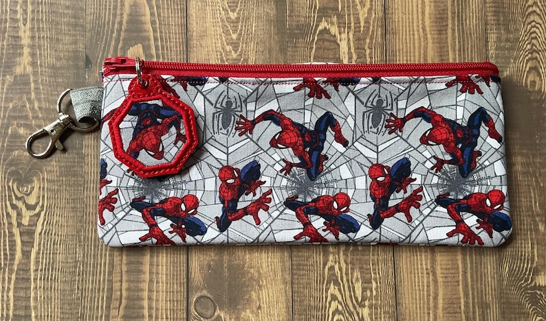 May include: A rectangular fabric pencil case featuring a repeating pattern of Spider-Man in red and blue suits against a gray spiderweb background. The case has a red zipper closure and a matching octagonal Spider-Man charm attached to a metal clip.