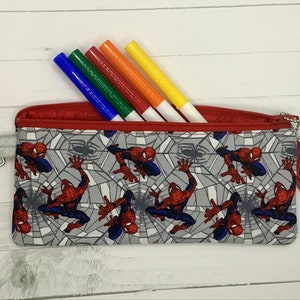 May include: A rectangular fabric pencil case featuring a repeating pattern of Spider-Man characters on a gray spiderweb background. The case has a red zipper closure, a silver metal lobster clasp on one side, and a matching hexagonal fabric charm.