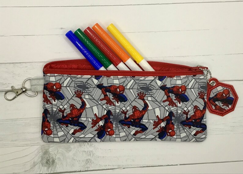 May include: A rectangular fabric pencil case featuring a repeating pattern of Spider-Man characters on a gray spiderweb background. The case has a red zipper closure, a silver metal lobster clasp on one side, and a matching hexagonal fabric charm.
