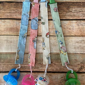 May include: Four baby pacifier clips with different colored fabric straps and pacifiers. The straps are blue, pink, cream, and green, each with a different pattern featuring bunnies and other illustrations. The pacifiers are blue, pink, and green.