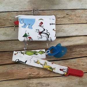 May include: A white fabric pacifier clip with a red clip and a white fabric pouch with a zipper. The pouch has a Dr. Seuss theme with characters like the Cat in the Hat and Thing 1 and Thing 2. The pouch is attached to the clip with a white fabric strap.