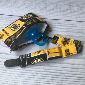 May include: A black, yellow, and white Bruins-themed pacifier case and clip. The zippered case holds a blue pacifier. The clip strap and case feature the Bruins logo and team name. The background is a light wood grain.
