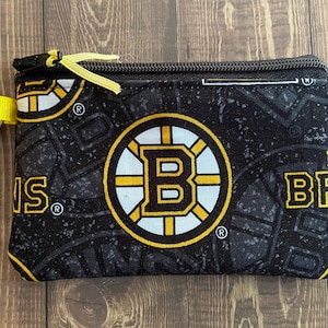 May include: Black coin purse with a Boston Bruins logo in yellow, white, and black. It has a yellow zip pull and a silver-coloured clip. The background pattern includes the team's logo and the word "BRUINS".