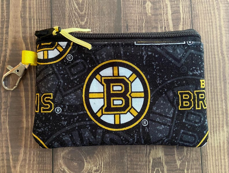 May include: Black coin purse with a Boston Bruins logo in yellow, white, and black. It has a yellow zip pull and a silver-coloured clip. The background pattern includes the team's logo and the word "BRUINS".