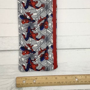 May include: A fabric pencil case featuring a repeating pattern of Spider-Man figures on a gray spiderweb background with a red zipper. A wooden ruler is placed along the bottom edge, showing the item is approximately 7.5 inches long.