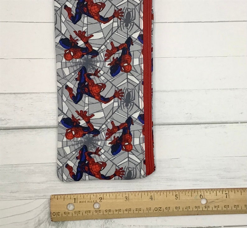 May include: A fabric pencil case featuring a repeating pattern of Spider-Man figures on a gray spiderweb background with a red zipper. A wooden ruler is placed along the bottom edge, showing the item is approximately 7.5 inches long.