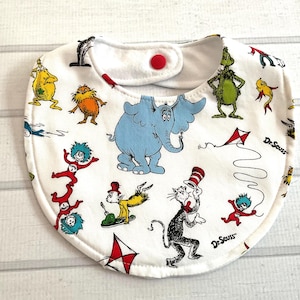 May include: White baby bib featuring colorful illustrations of characters from Dr. Seuss books. The bib has a red snap closure and is decorated with images of the Cat in the Hat, the Grinch, and Horton the Elephant.