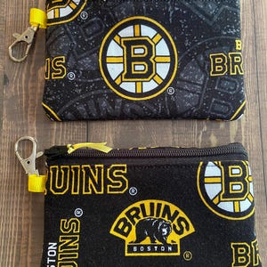 May include: Two black zippered pouches with yellow accents and Boston Bruins logos. The pouches feature the team's logo, a bear, and the words "Bruins" and "Boston" in yellow and white. Each pouch has a yellow wrist strap and a silver clasp.