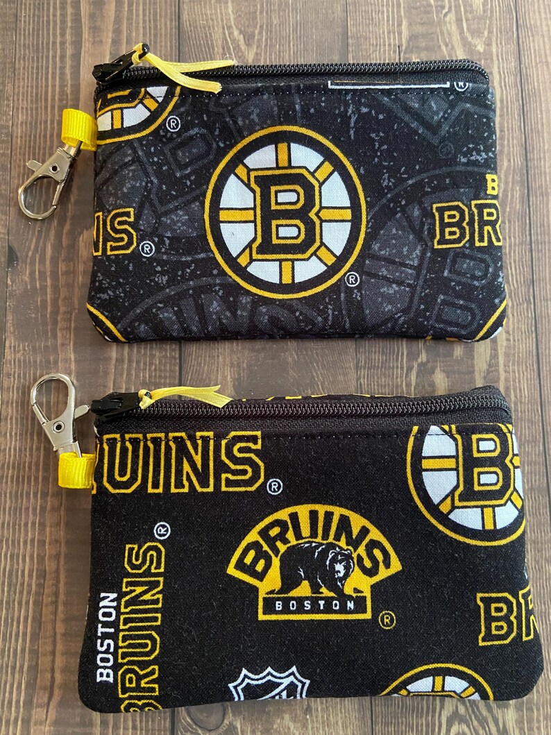 May include: Two black zippered pouches with yellow accents and Boston Bruins logos. The pouches feature the team's logo, a bear, and the words "Bruins" and "Boston" in yellow and white. Each pouch has a yellow wrist strap and a silver clasp.