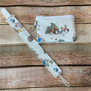 May include: A light-colored fabric lanyard and small zippered pouch featuring illustrations of rabbits and other characters. The lanyard has a white plastic clip and a loop at the end. The pouch has a silver zipper and a silver clasp.