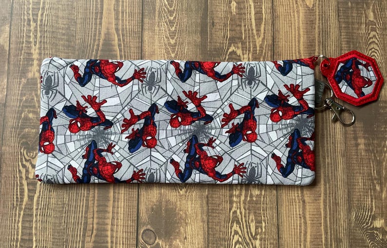 May include: A rectangular fabric pencil case featuring a repeating pattern of Spider-Man in red and blue suits against a gray spiderweb background. A matching hexagonal keychain with a red border and a metal lobster clasp is attached to the side.