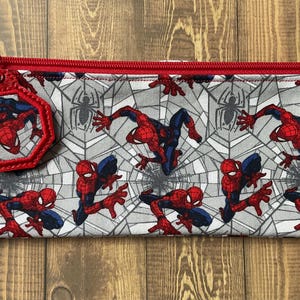May include: A rectangular fabric pencil case featuring a repeating pattern of the superhero Spider-Man in red and blue against a gray spiderweb background. The case has a red zipper closure and a matching octagonal charm attached to the zipper pull.