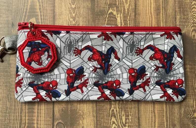 May include: A rectangular fabric pencil case featuring a repeating pattern of the superhero Spider-Man in red and blue against a gray spiderweb background. The case has a red zipper closure and a matching octagonal charm attached to the zipper pull.