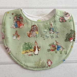 May include: A green baby bib with a white lining and snap closure. The bib features a whimsical print of rabbits, an owl, and other woodland creatures in a garden setting. The bib is made of soft fabric.