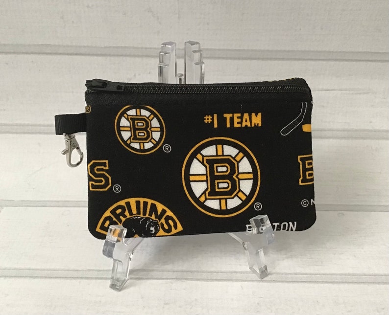 May include: Black zippered pouch with a repeating pattern of the Boston Bruins logo, the words "#1 TEAM" and "BOSTON". The pouch has a black zipper and a silver clip.