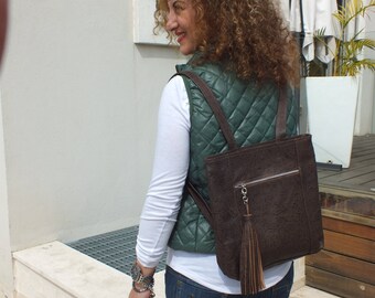 Può includere: Uno zaino in pelle marrone con un lungo pendente a nappa che pende dalla cerniera. Lo zaino ha un gilet trapuntato verde sopra.