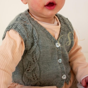 May include: A knitted baby vest in a soft, muted green. The vest has a V-neck, button closure, and a delicate leaf pattern on the front. The buttons are heart-shaped. The baby is wearing a peach-coloured long-sleeved shirt underneath.