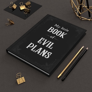 May include: A black notebook with the text "My little BOOK of EVIL PLANS" in white. Gold and black pencils and gold binder clips are arranged around the notebook on a dark gray surface. Decorative office supplies.