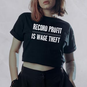 May include: Black t-shirt with the white text "RECORD PROFIT IS WAGE THEFT". The shirt has short sleeves and a rolled hem. The person is wearing black pants.