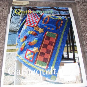 May include: A blue quilt with colorful geometric shapes, including squares, stars, and rings. The quilt has a checkerboard pattern and the word "Gamequilt" is visible. The quilt is by Rhode Island.