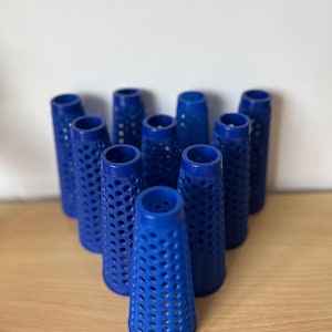 Peut inclure: Un ensemble de bigoudis bleus en forme de cône avec un motif perforé. Les bigoudis sont disposés en triangle, les extrémités ouvertes vers le haut. Le fond est un mur blanc uni et une surface en bois.