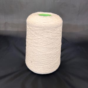 Peut inclure: Un grand cône de fil blanc cassé, étroitement enroulé avec une texture de fil visible. Le dessus du cône a un centre vert. Le fil est posé sur un fond sombre.