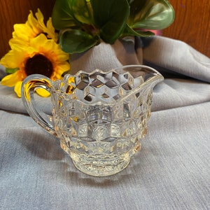 May include: A clear glass creamer with a textured, diamond-shaped pattern. The creamer has a handle and a spout.