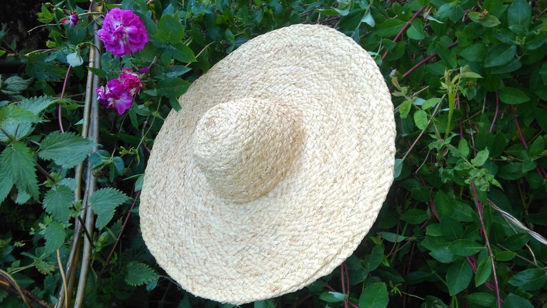 May include: A large, wide-brimmed straw hat with a woven texture. The hat is light beige and is sitting on a bed of green leaves.