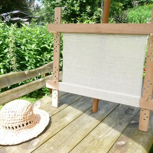 May include: A wooden embroidery hoop with a beige linen fabric stretched across it. The hoop is standing on a wooden table with a straw hat on the table.