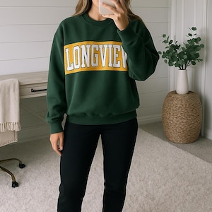 May include: A dark green sweatshirt with the word "LONGVIEW" in white block letters on a yellow band across the chest. The sweatshirt is paired with black joggers and white sneakers. The outfit is casual and comfortable.