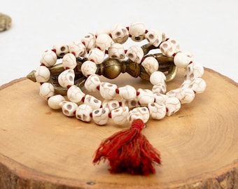 May include: White skull bead mala with a red tassel, resting on a wooden slice. The mala is made of bone and has a brass charm.