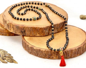 May include: Black beaded necklace with a skull charm and red tassel. The necklace is displayed on a wooden slice.