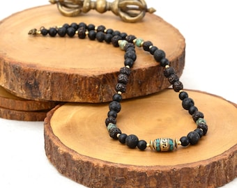 May include: A black beaded necklace with gold accents and a turquoise and gold cylindrical bead. The necklace is displayed on a wooden slice with a gold-colored metal object in the background. The necklace is made of black lava rock beads.
