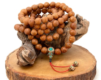 May include: A strand of light brown prayer beads with a decorative pendant featuring turquoise and red accents. The beads are displayed on a wooden base with a natural, rustic appearance.