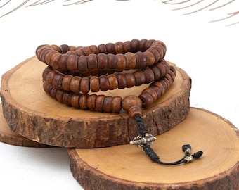 May include: A brown wooden mala bead necklace with a silver charm and black cord. The necklace is displayed on two wooden slices.