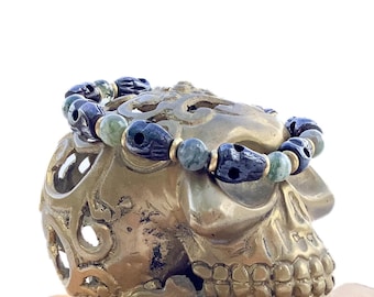 May include: A black and green beaded bracelet with skull-shaped beads, resting on a gold skull sculpture on a stack of wooden coasters.