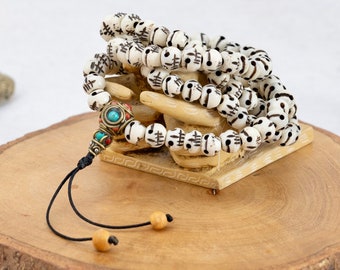 May include: White prayer beads with black markings and a turquoise and red tassel. The beads are arranged on a wooden hand sculpture.