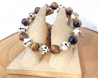 May include: A beaded bracelet featuring a mix of brown tiger eye, black, and cream-colored beads with a skull design. The bracelet is displayed on a carved, beige hand-shaped stand, set on a wooden slice.