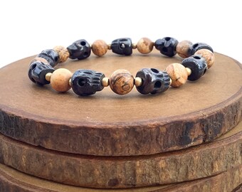 May include: A black and brown skull bead bracelet with gold spacers. The bracelet is displayed on a stack of wooden rounds.