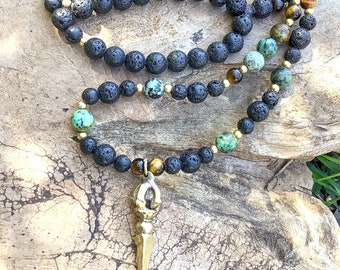 May include: A black lava rock, turquoise, and tiger's eye beaded necklace with a gold pendant. The necklace is on a wooden surface.