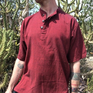Puede incluir: Una camisa de manga corta de color burdeos con cuello mandarín y cierre de botones. La camisa tiene un corte holgado y está hecha de una tela suave y ligera.