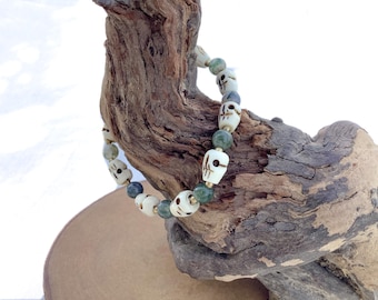 May include: A white skull bead bracelet with green gemstone beads. The bracelet is displayed on a piece of driftwood.