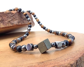 May include: A beaded necklace with brown, black, and silver beads. A square-shaped, dark green stone is in the center of the necklace.